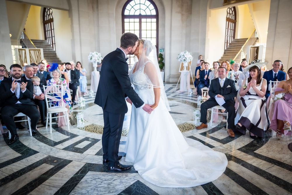 Bride and groom share first kiss at elegant wedding ceremony in grand hall with guests applauding.