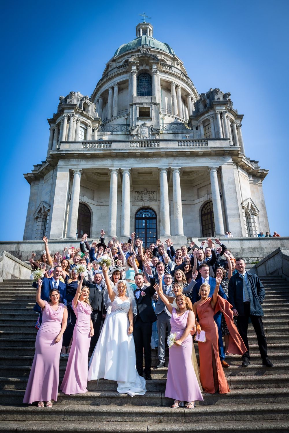 Wedding party celebrating on steps of grand domed building with bridesmaids in pink dresses under blue sky.