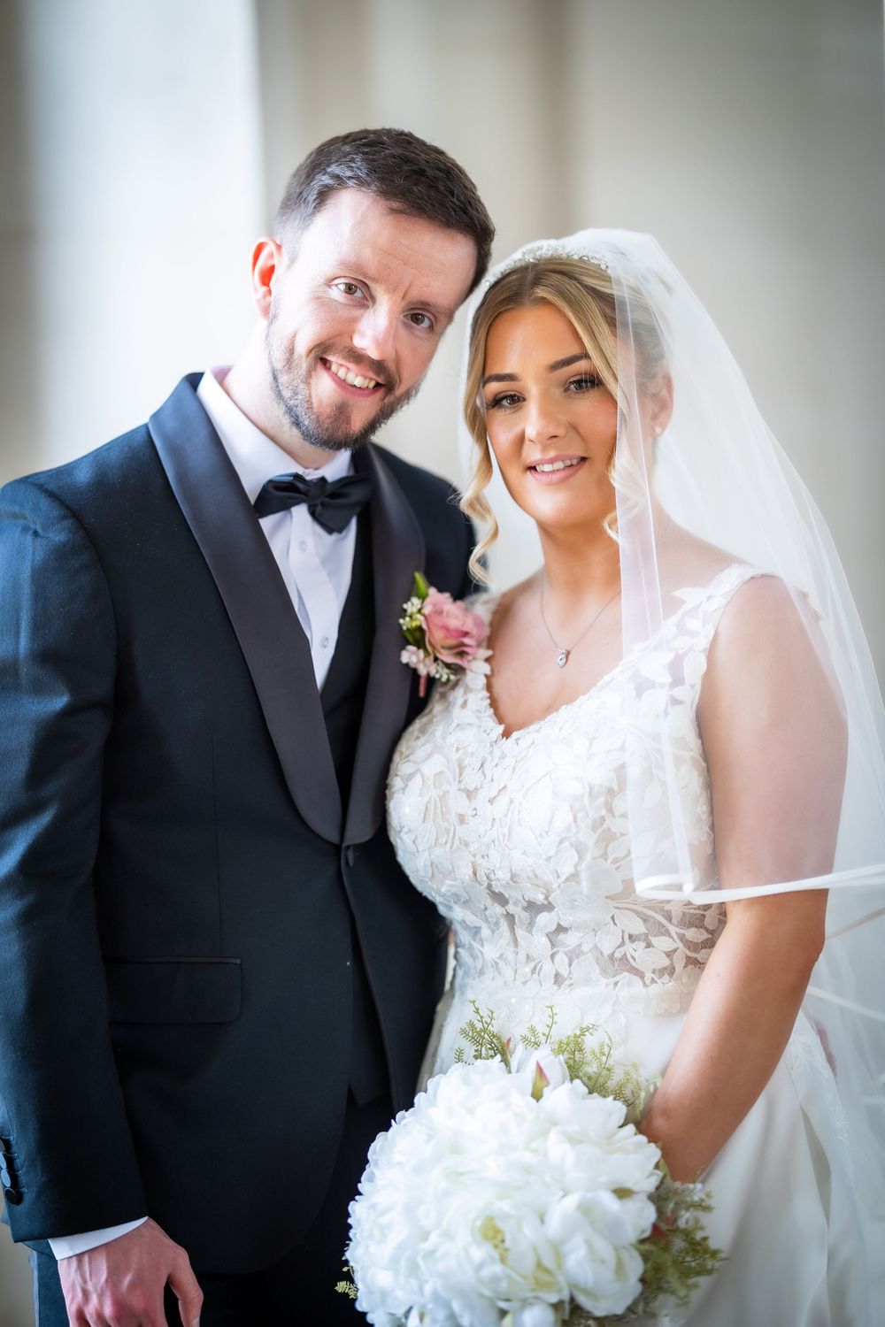 Smiling bride and groom posing together on their wedding day, bride holding white bouquet wearing veil, groom in black tuxedo.