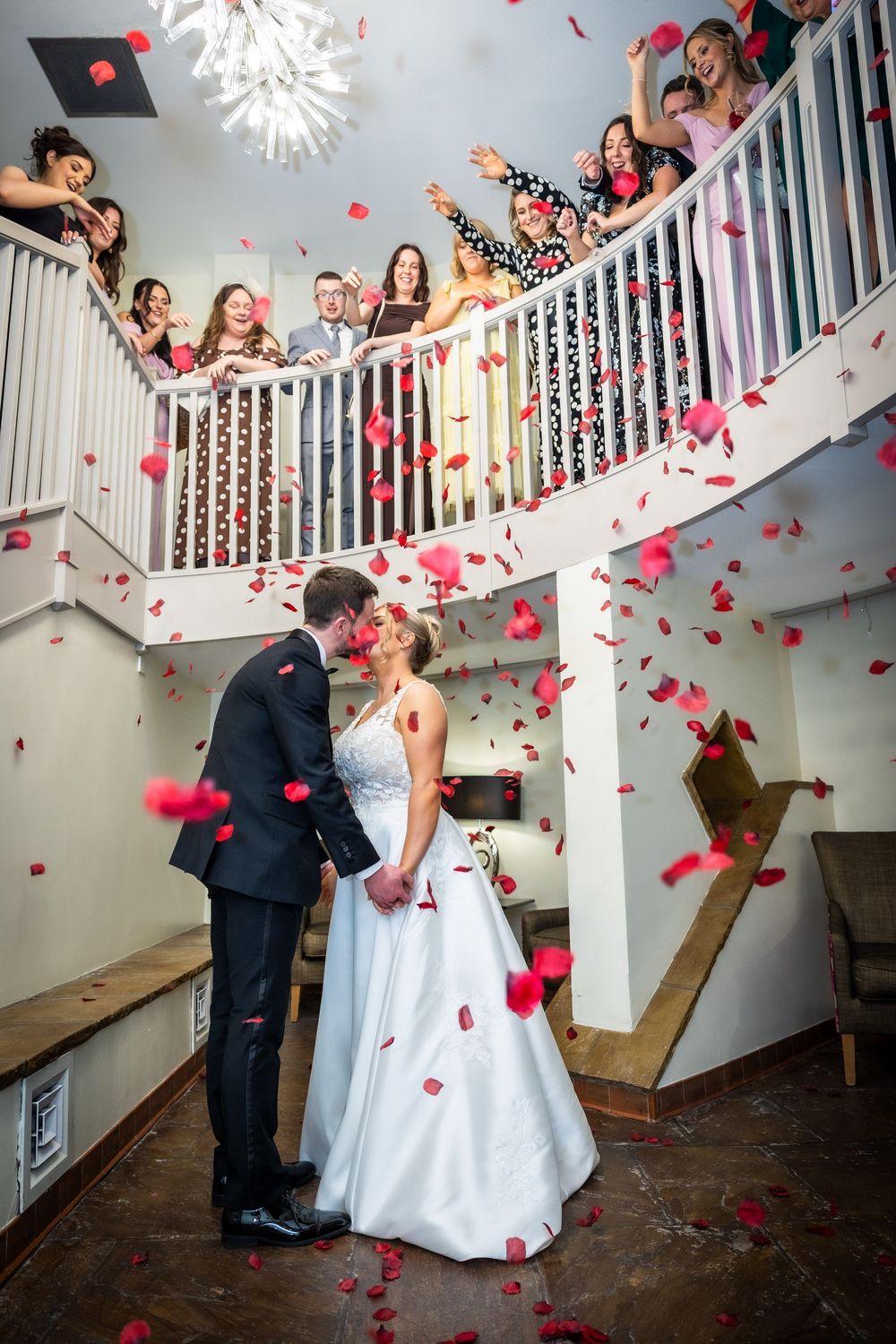Bride and groom kissing under falling red rose petals as guests celebrate from balcony above staircase.