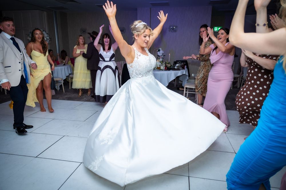 Bride in white ball gown twirling on dance floor surrounded by wedding guests clapping and celebrating.