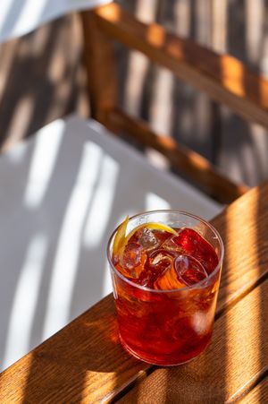 Red cocktail with ice and lemon slice in rocks glass on sunny wooden table.