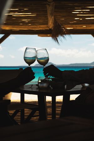 Two hands clinking wine glasses on a beach table with turquoise ocean views under a thatched roof.