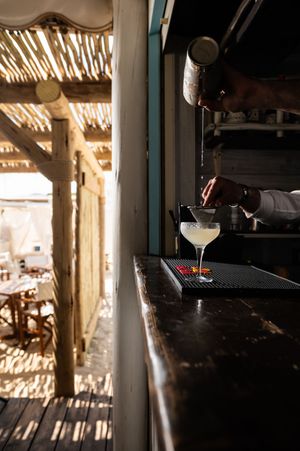 Bartender straining a cocktail into a coupe glass at a rustic beachside bar with thatched roof.