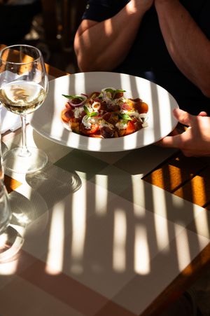 A colorful gourmet salad in a white bowl served with a glass of white wine on a sunlit restaurant table.