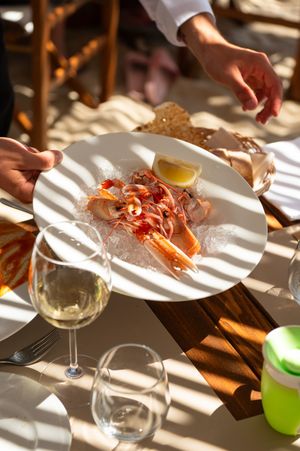 Hands serving a white plate of fresh shrimp on ice with lemon wedge alongside a glass of white wine.