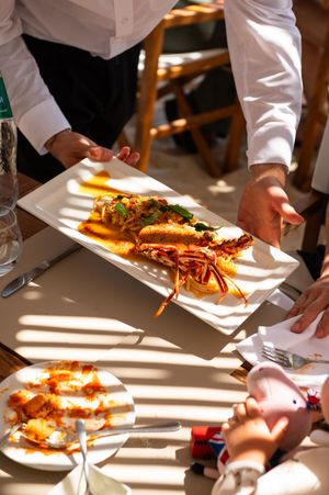 Waiter serving a grilled lobster dish on a white rectangular plate at a sunlit restaurant table.