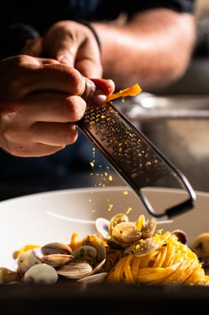 Chef grating lemon zest over a plate of linguine alle vongole with fresh clams in a restaurant kitchen.