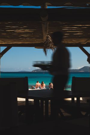 Silhouette of a waiter carrying a tray at a beachside bar with turquoise ocean views and sunbathers on the shore.