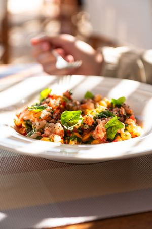 A white plate of pasta with tomato sauce, ground meat, and fresh basil leaves served in a sunlit restaurant.