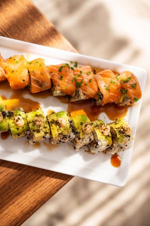 Two rows of fresh sushi rolls on a white plate, topped with salmon, avocado, sesame seeds, and teriyaki sauce.