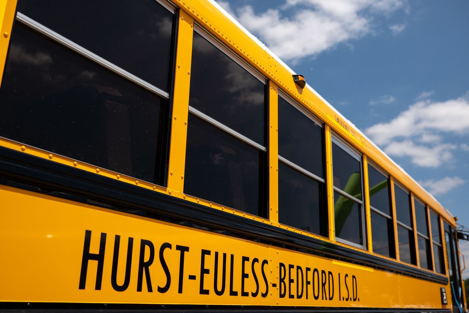 Yellow Hurst-Euless-Bedford ISD school bus parked outdoors under a partly cloudy blue sky.