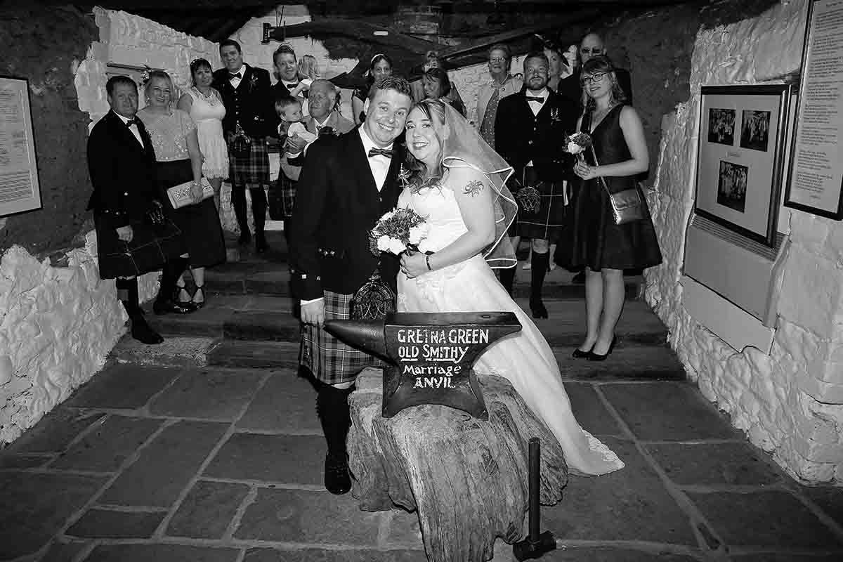 Black and white wedding photo of a couple in Scottish attire at Gretna Green, surrounded by guests in a stone building.