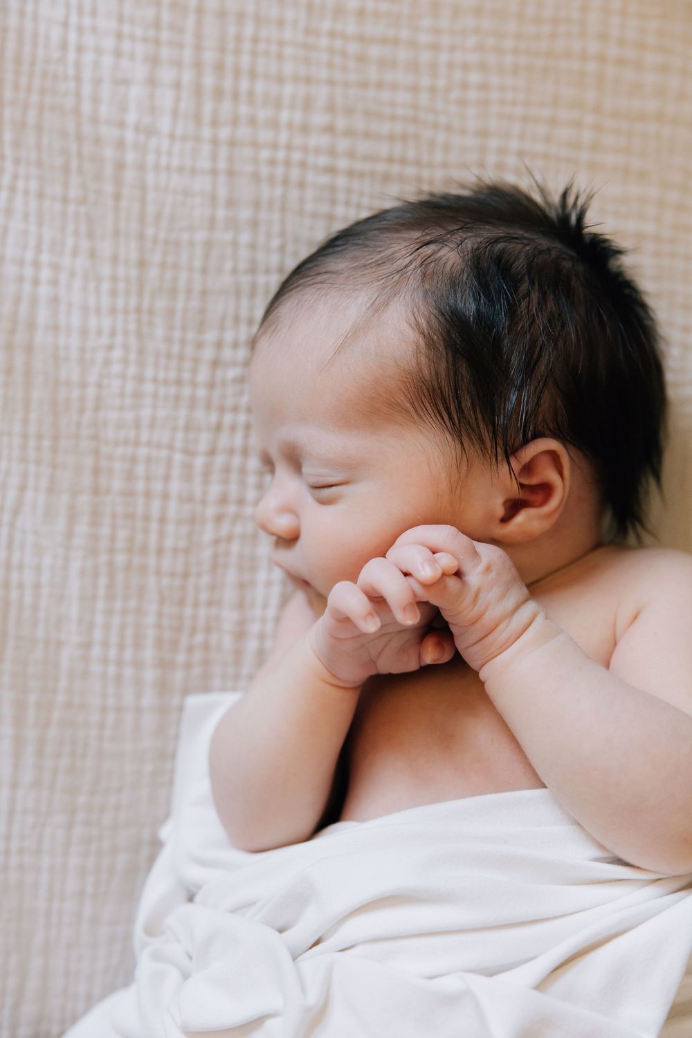 Peaceful newborn baby sleeping with hands near face, wrapped in soft white blanket.