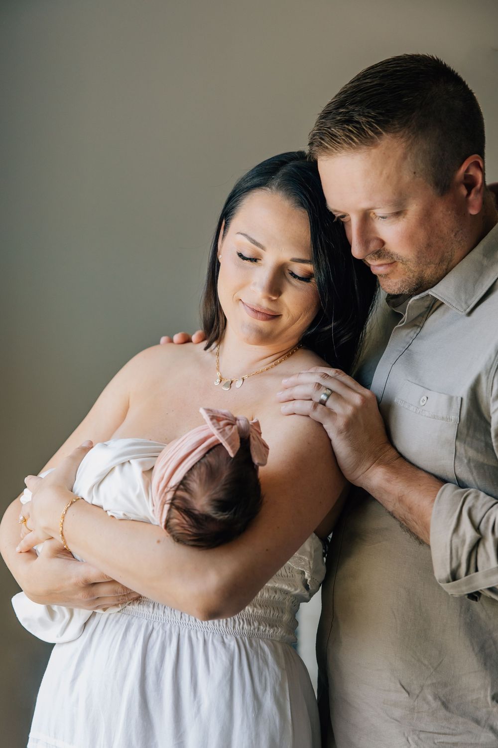 A couple tenderly gazes at their newborn baby, mother in white dress holding infant with pink headband, father in gray shirt.