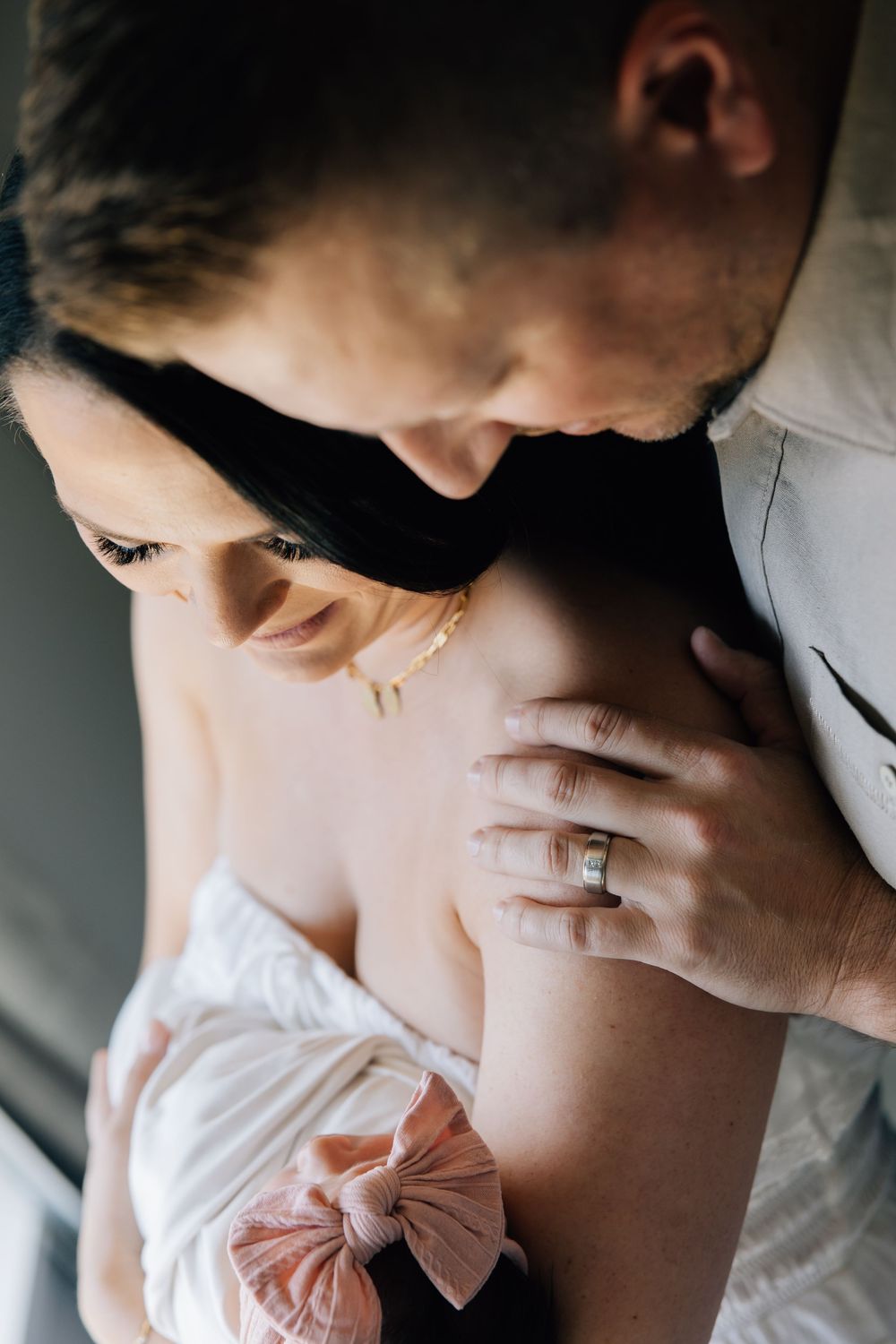 A couple shares an intimate moment, the man gently kissing the woman's head as she holds a newborn with a pink bow.