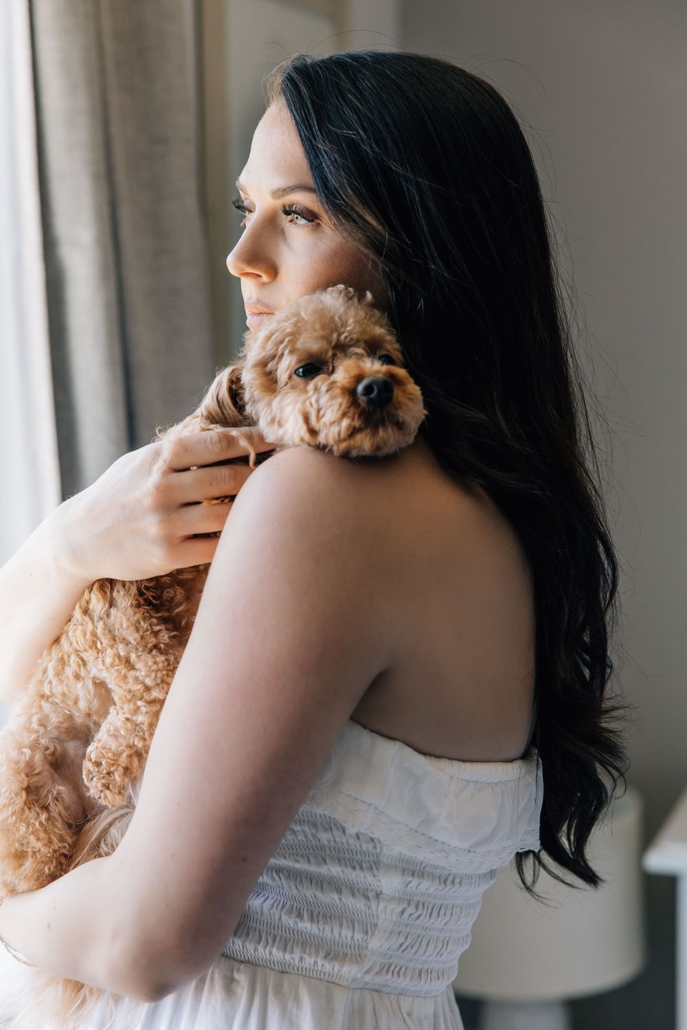 Dark-haired woman in white strapless dress holds a fluffy brown Cavapoo puppy near a bright window.
