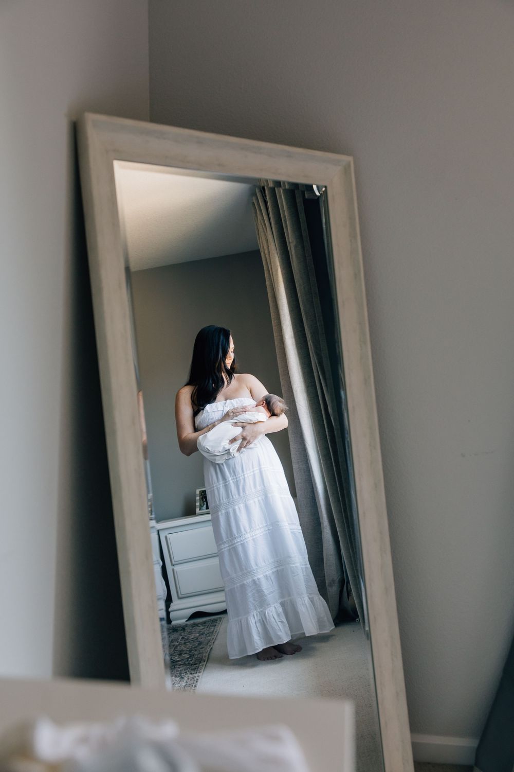 Woman in white dress holding newborn baby reflected in full-length mirror in nursery room.