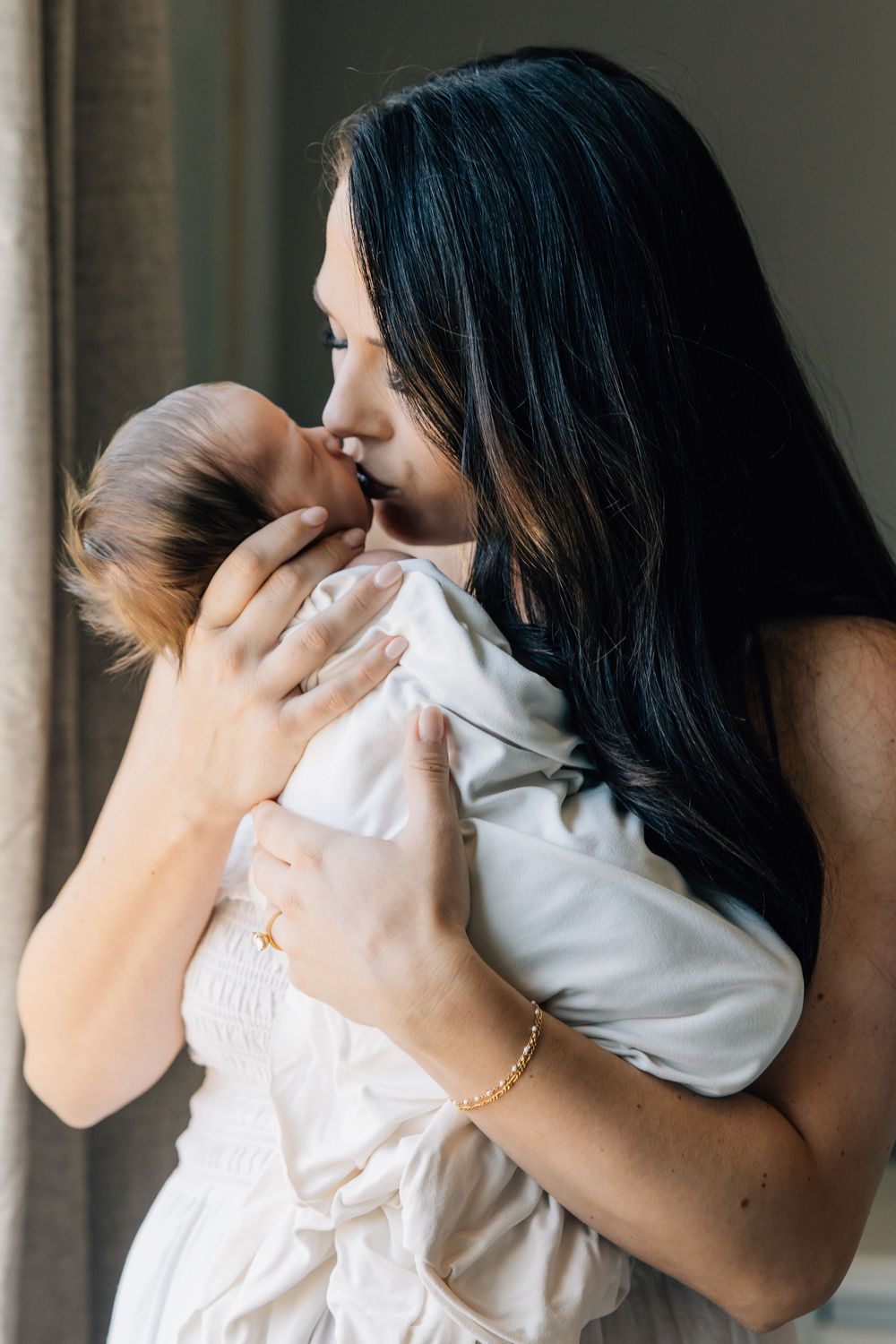 A dark-haired mother tenderly kisses her swaddled newborn baby while standing near a window.