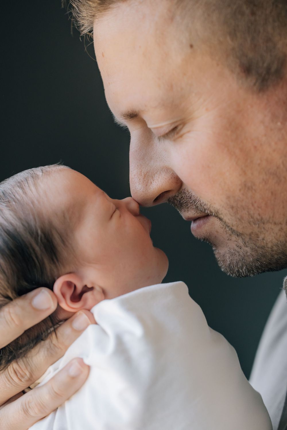 Father tenderly touches noses with his newborn baby, cradling the infant gently in his hands against a dark background.