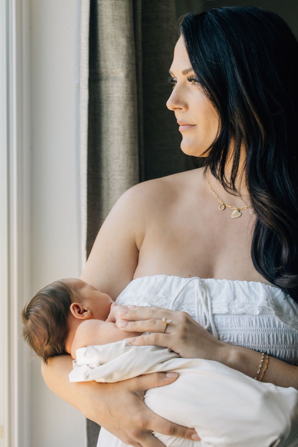 Dark-haired woman in white off-shoulder top tenderly holds sleeping newborn baby near a sunlit window.