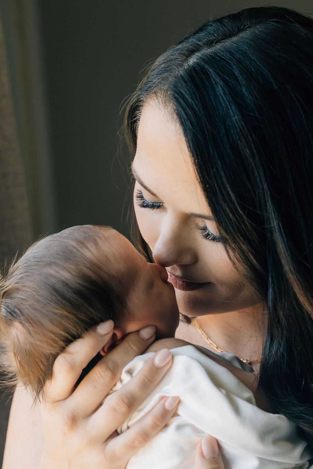 A dark-haired mother lovingly nuzzles her newborn baby in a softly lit room.