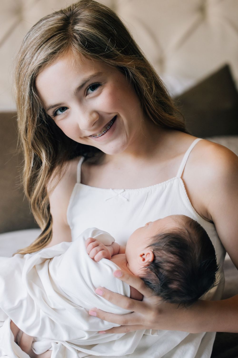 Smiling teenage girl with braces holds a sleeping newborn baby wrapped in white blanket while sitting on a couch.