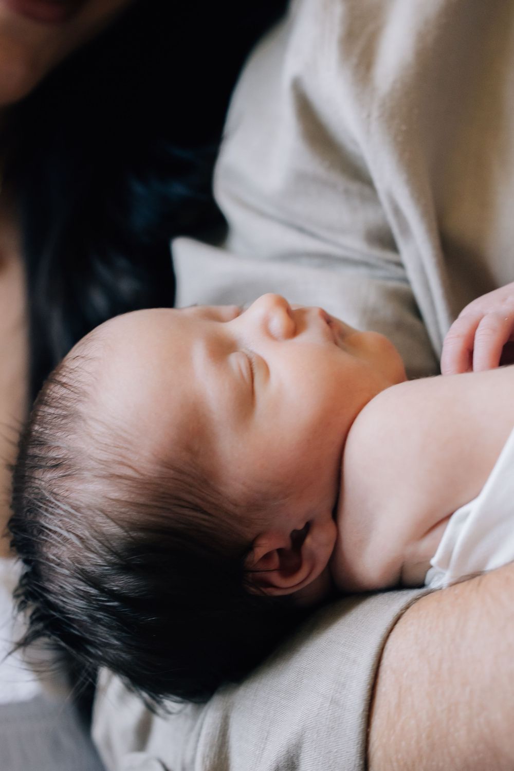 A peaceful newborn baby with dark hair sleeping soundly in a parent's arms, wearing a white onesie.