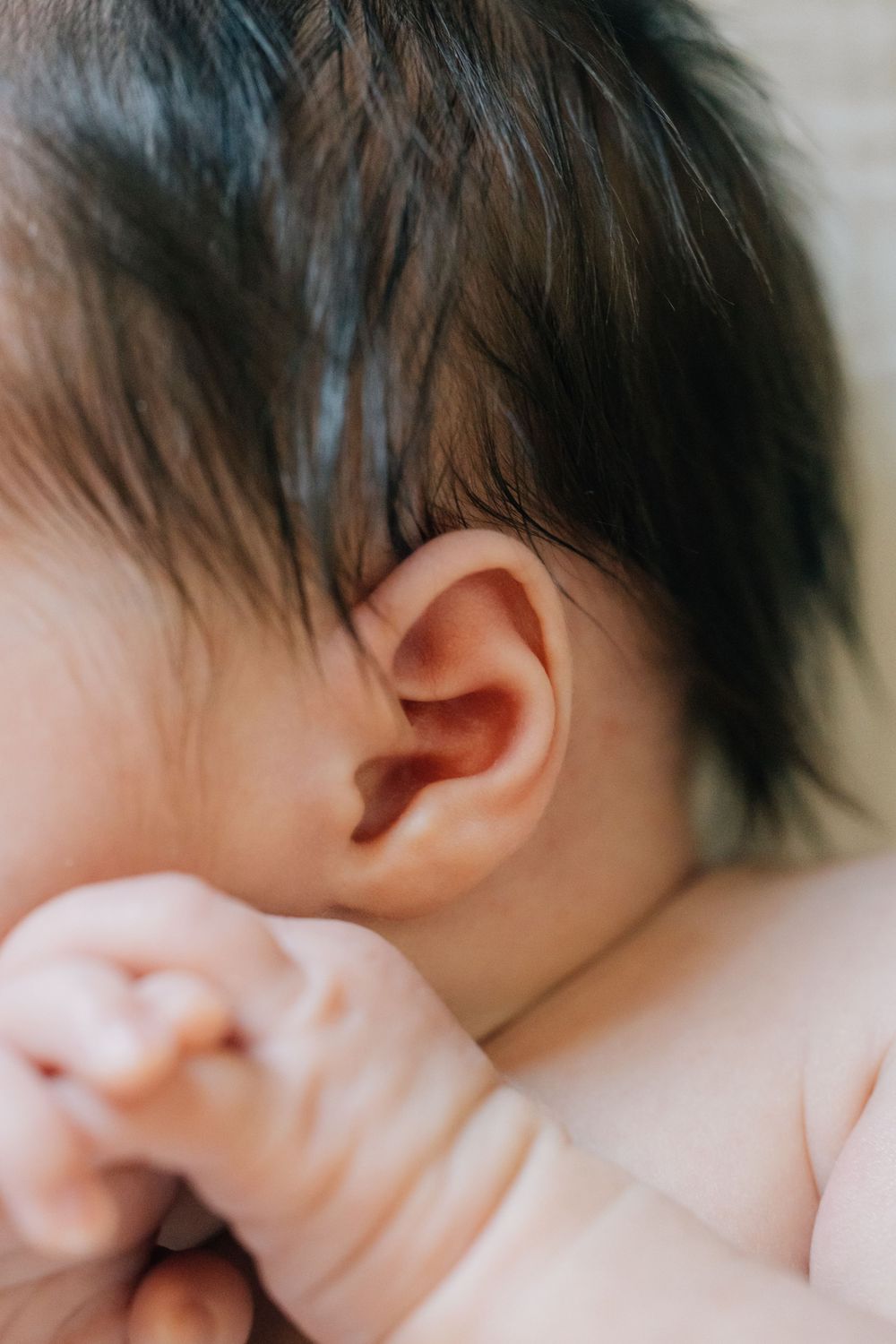Close-up of a newborn baby's ear with dark hair visible, tiny hand nearby.