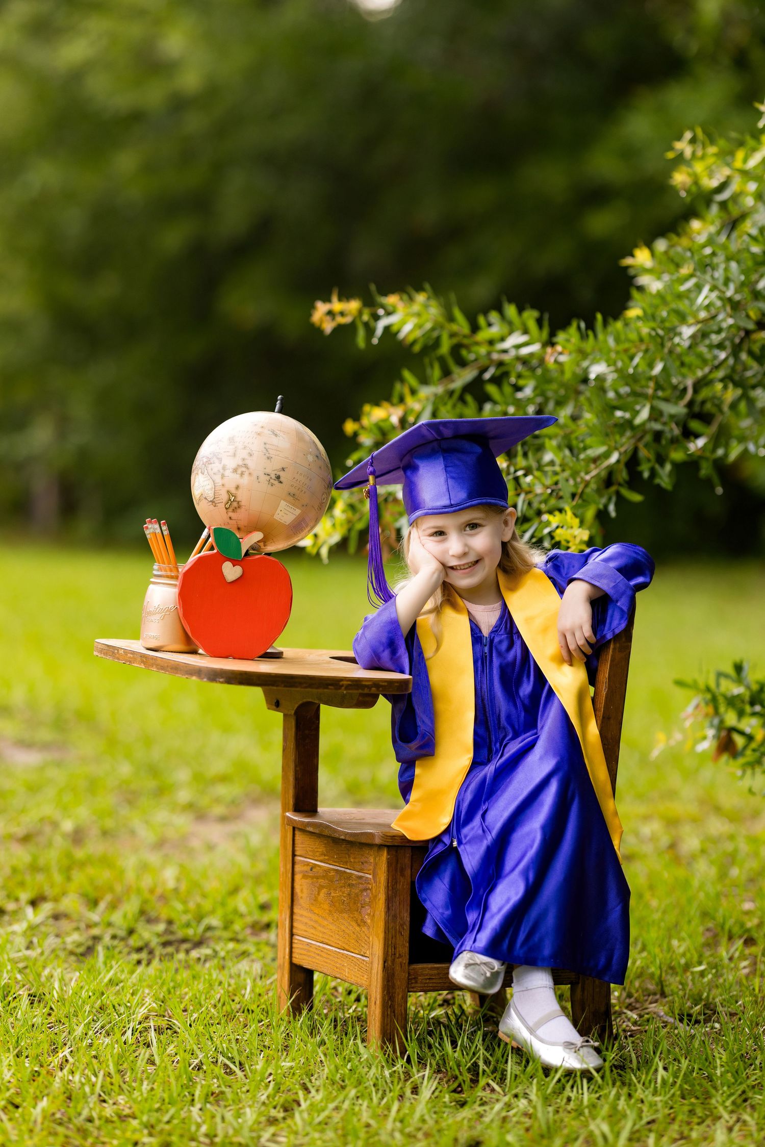 Young girl in blue graduation cap and gown sits at small wooden desk outdoors with globe and apple decorations.