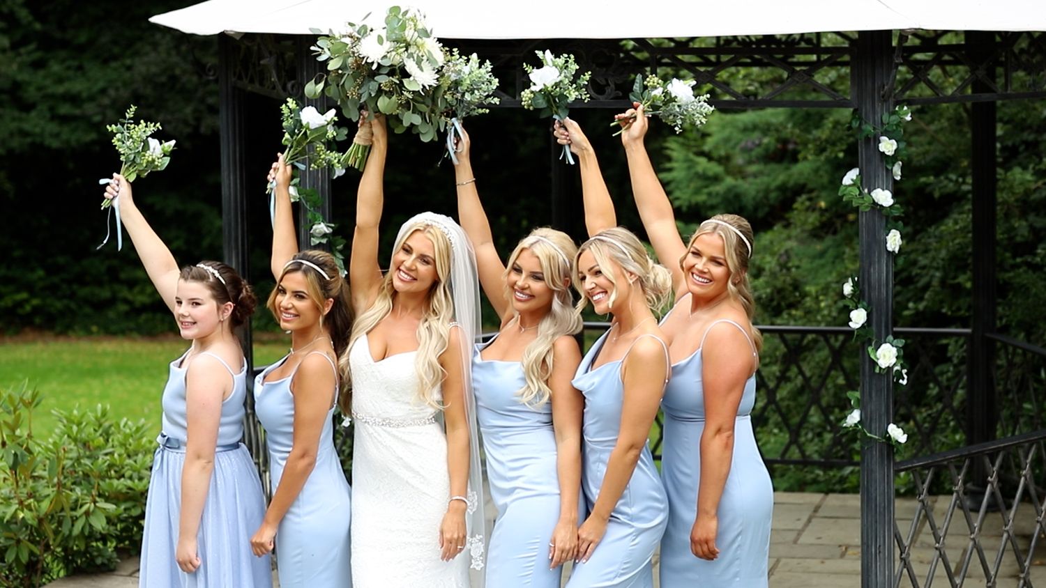 Bridesmaids in light blue dresses joyfully raise their bouquets while celebrating outdoors.