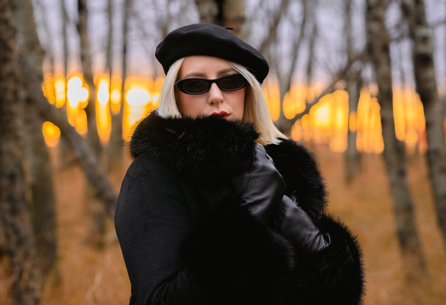 Figure in black winter attire with sunglasses stands in autumn forest with glowing orange sunset in background.