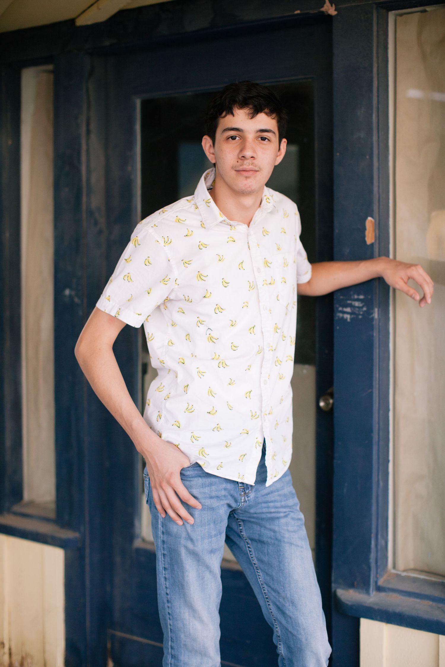 Casual pose in doorway wearing white patterned shirt and blue jeans.