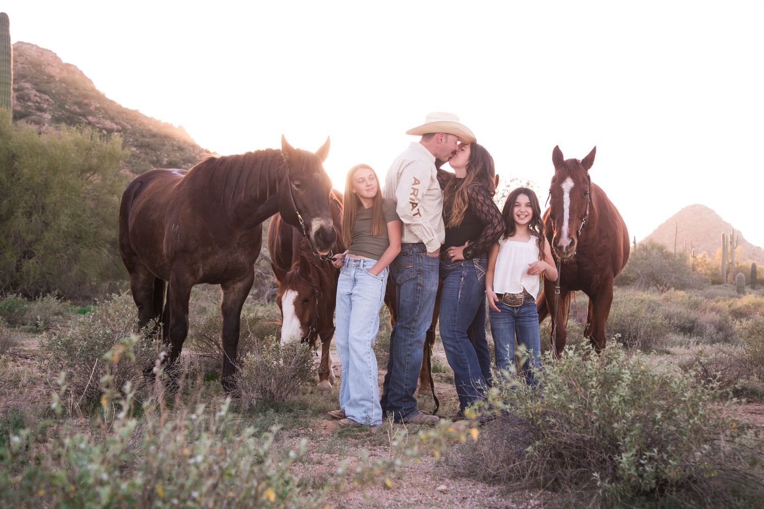 Family with two daughters poses with three brown horses at golden hour in desert landscape with cacti.