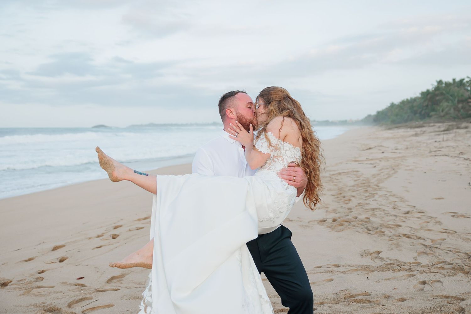 A bride and groom embrace on a beach, with the ocean and rocky coastline in the background.