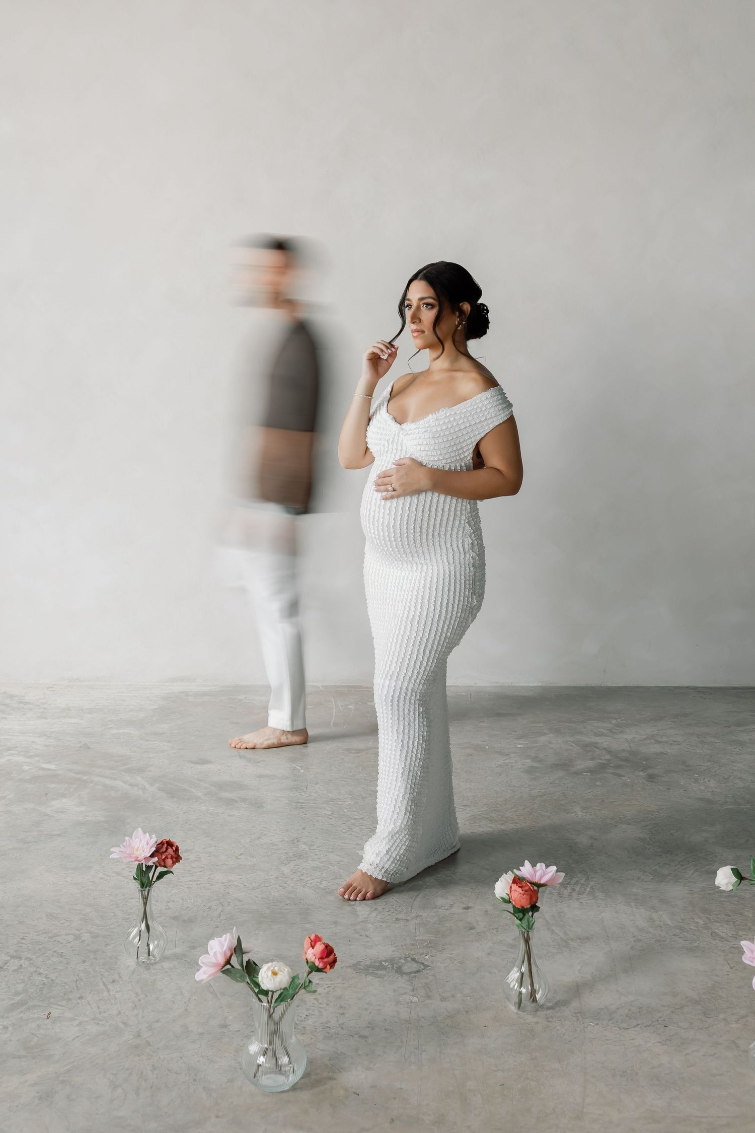 A woman in a long white dress stands in a room with a man in the background.