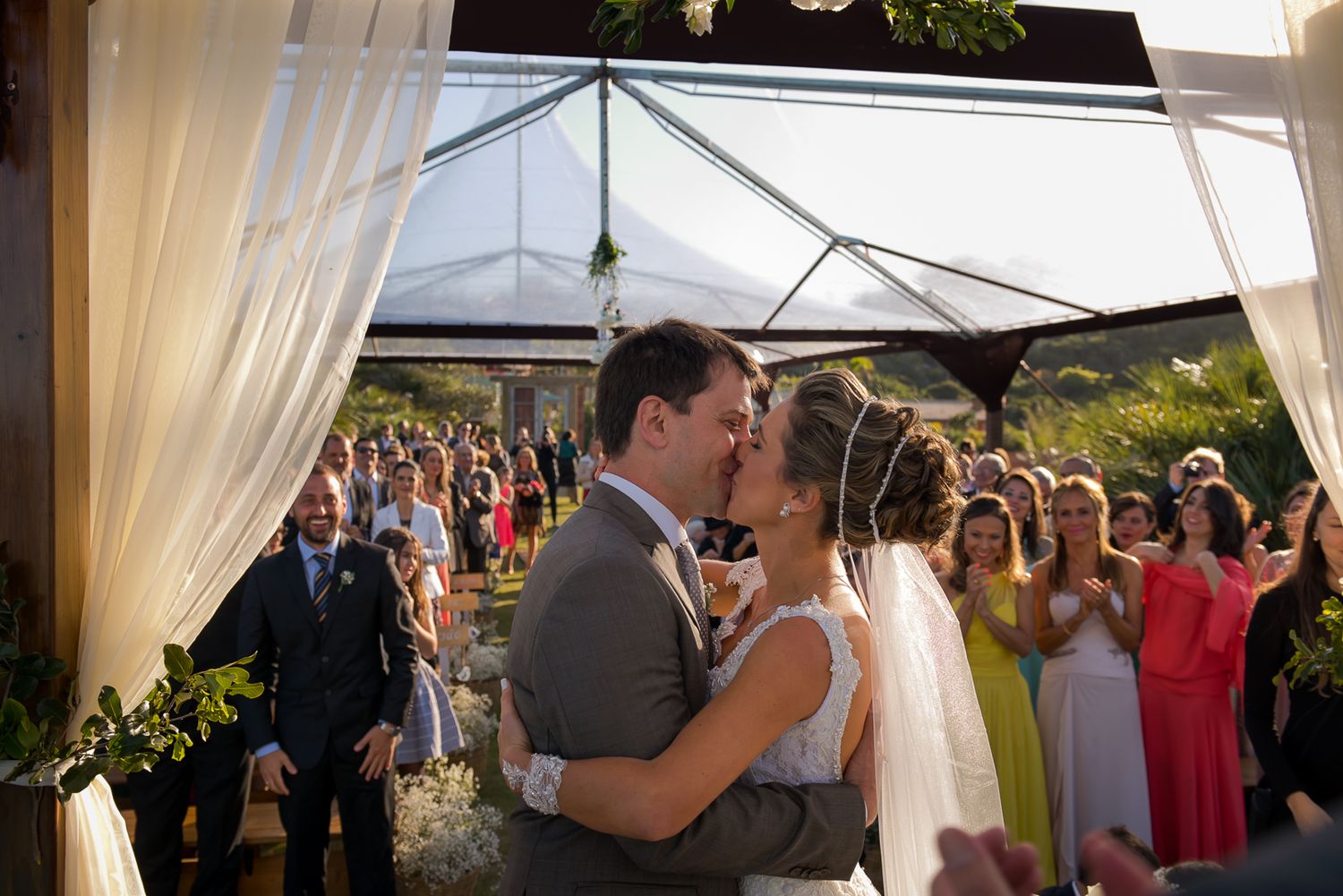 Newlyweds share their first kiss under a white draped tent as guests watch and celebrate.