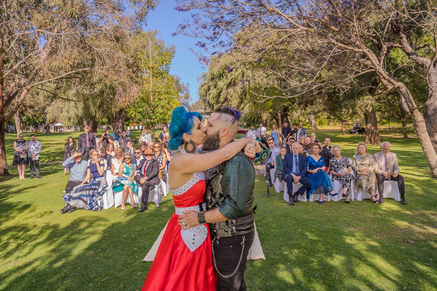 A couple shares their first dance outdoors during a wedding ceremony with guests seated in the background on a sunny day.
