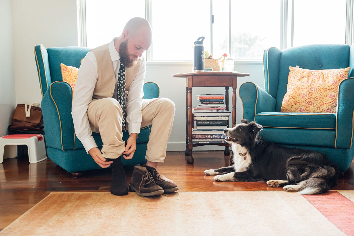 A person in beige clothing ties their shoes in a living room while a black and white dog rests by a teal armchair.