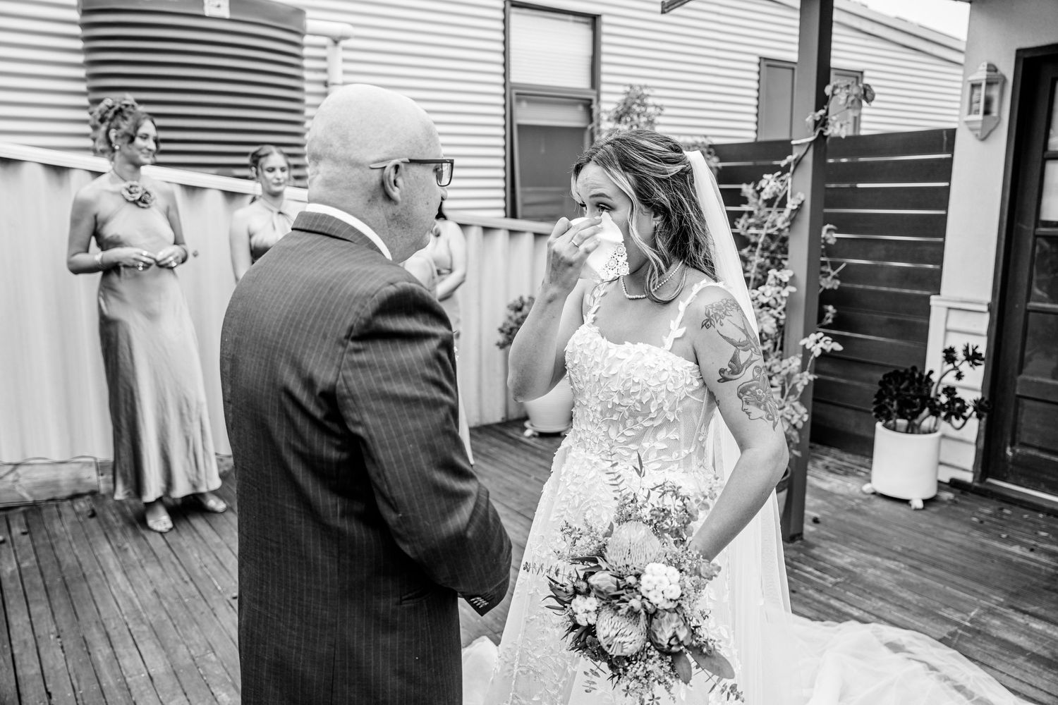 A touching wedding moment in black and white captured on a wooden deck with elegant floral bouquet details.