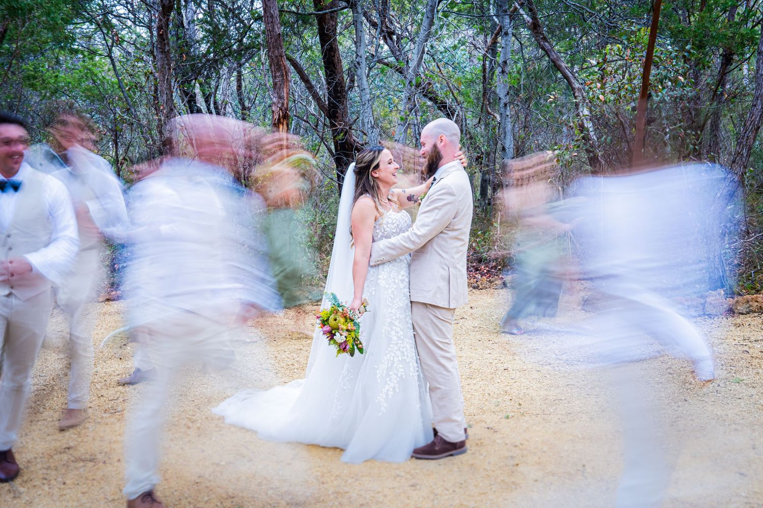 A couple shares a romantic moment at their outdoor wedding while guests move in motion blur around them on a forest path.