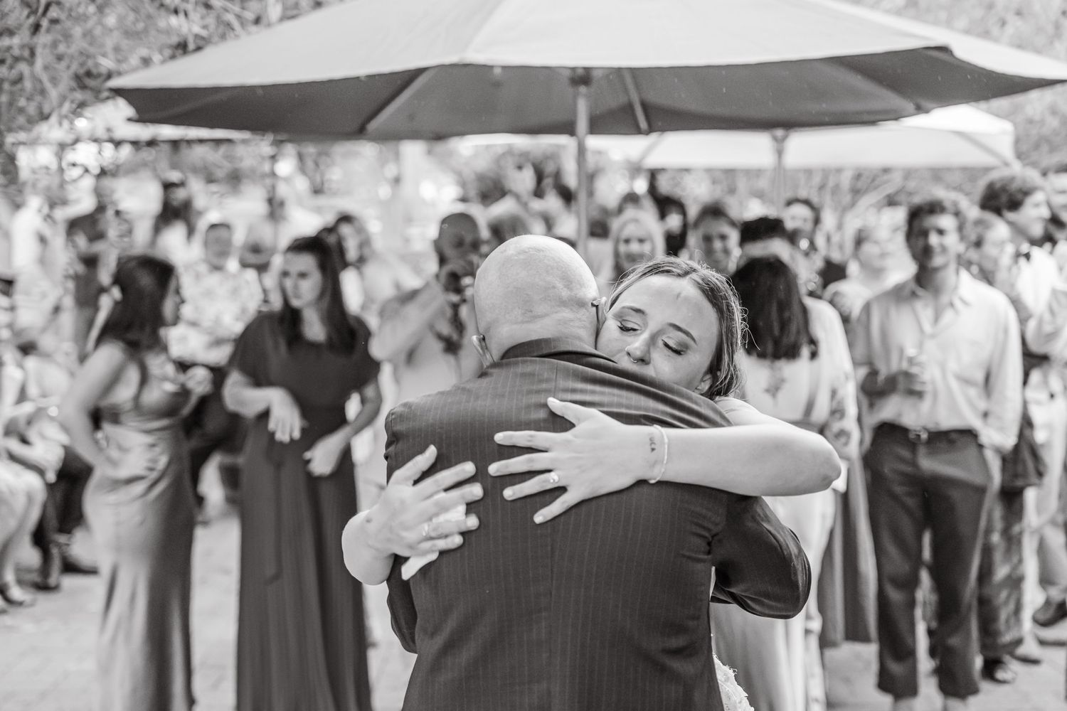 Black and white photo of two people embracing at an outdoor event with guests and umbrellas in the background.