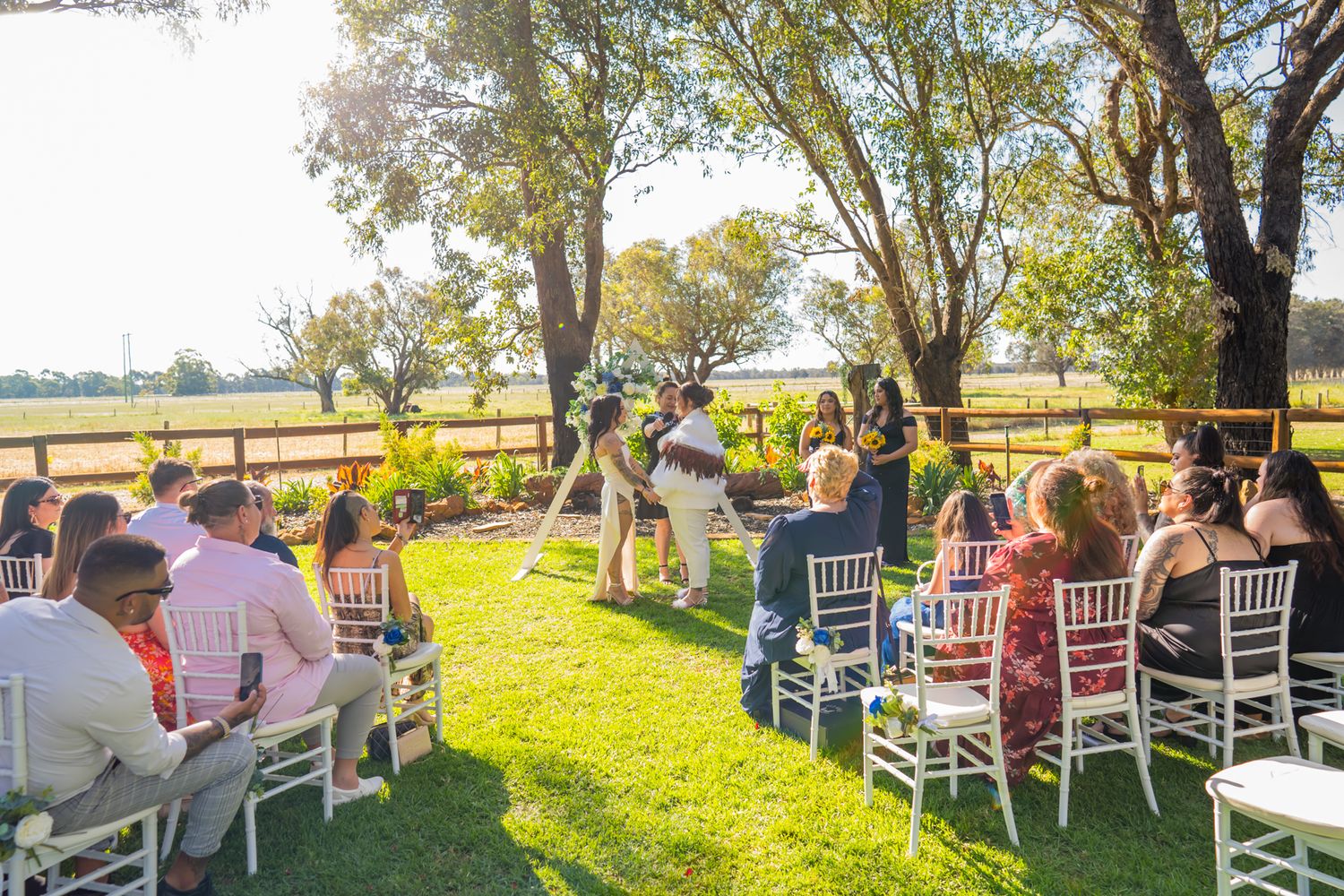 Outdoor wedding ceremony setup with white chairs under tall trees on a sunny afternoon.