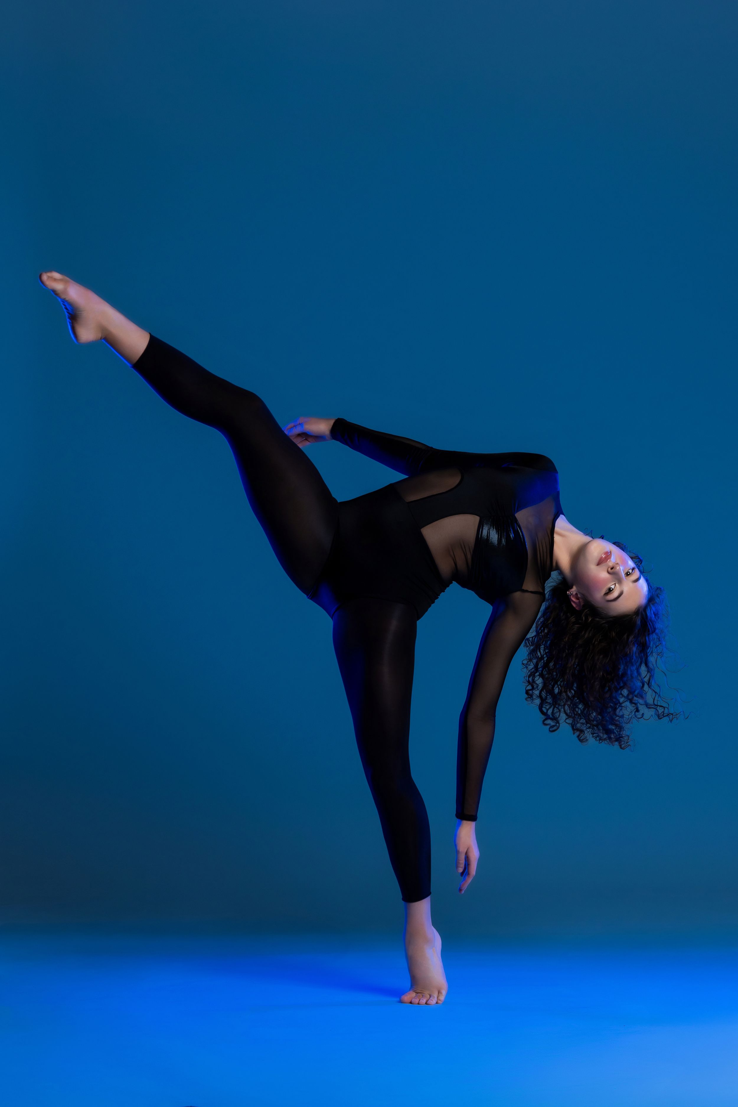 Dancer in black athletic wear performs a dynamic high kick pose against a blue backdrop with dramatic lighting.