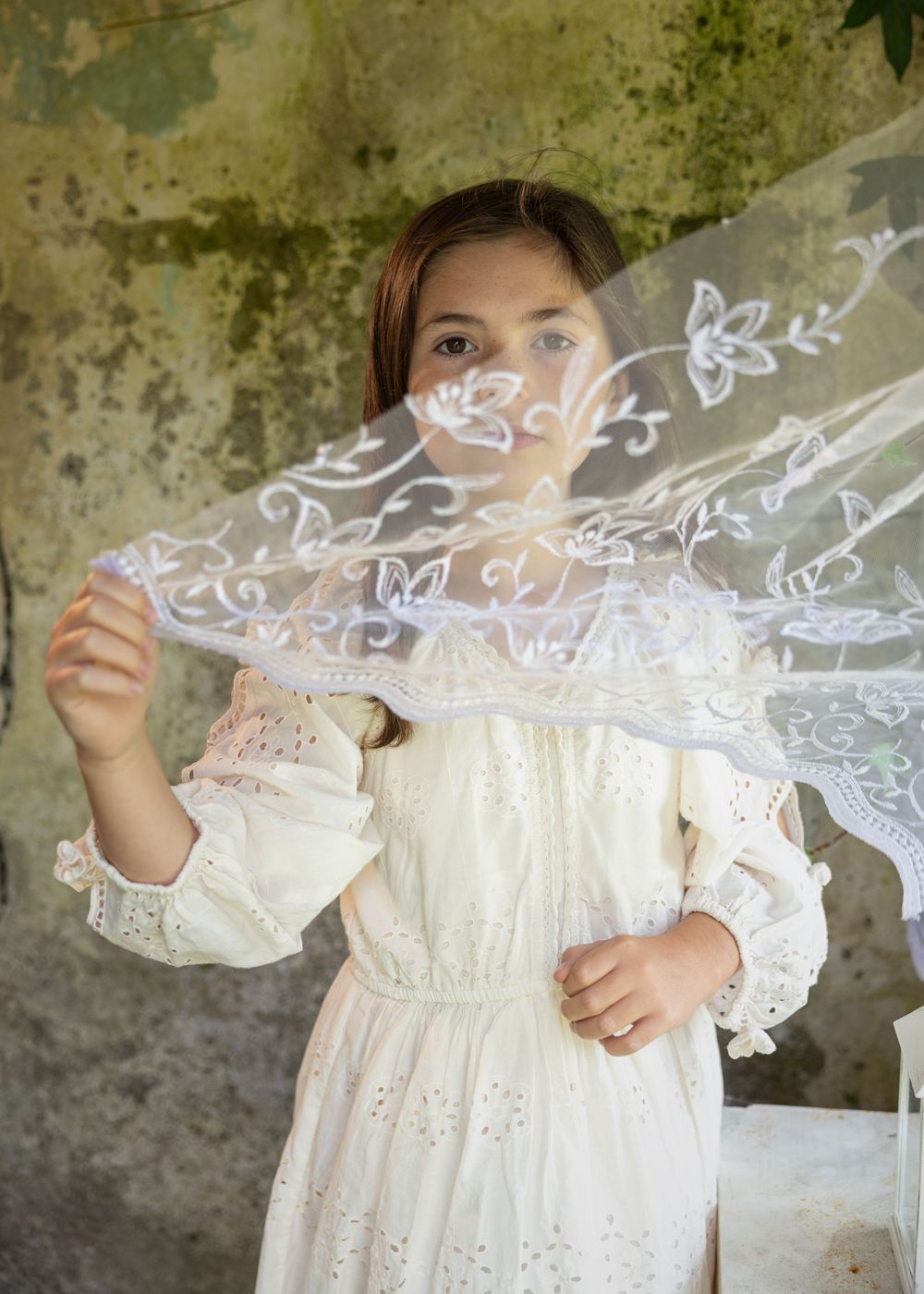 A delicate white lace dress with intricate embroidery details is displayed against a weathered stone wall background.