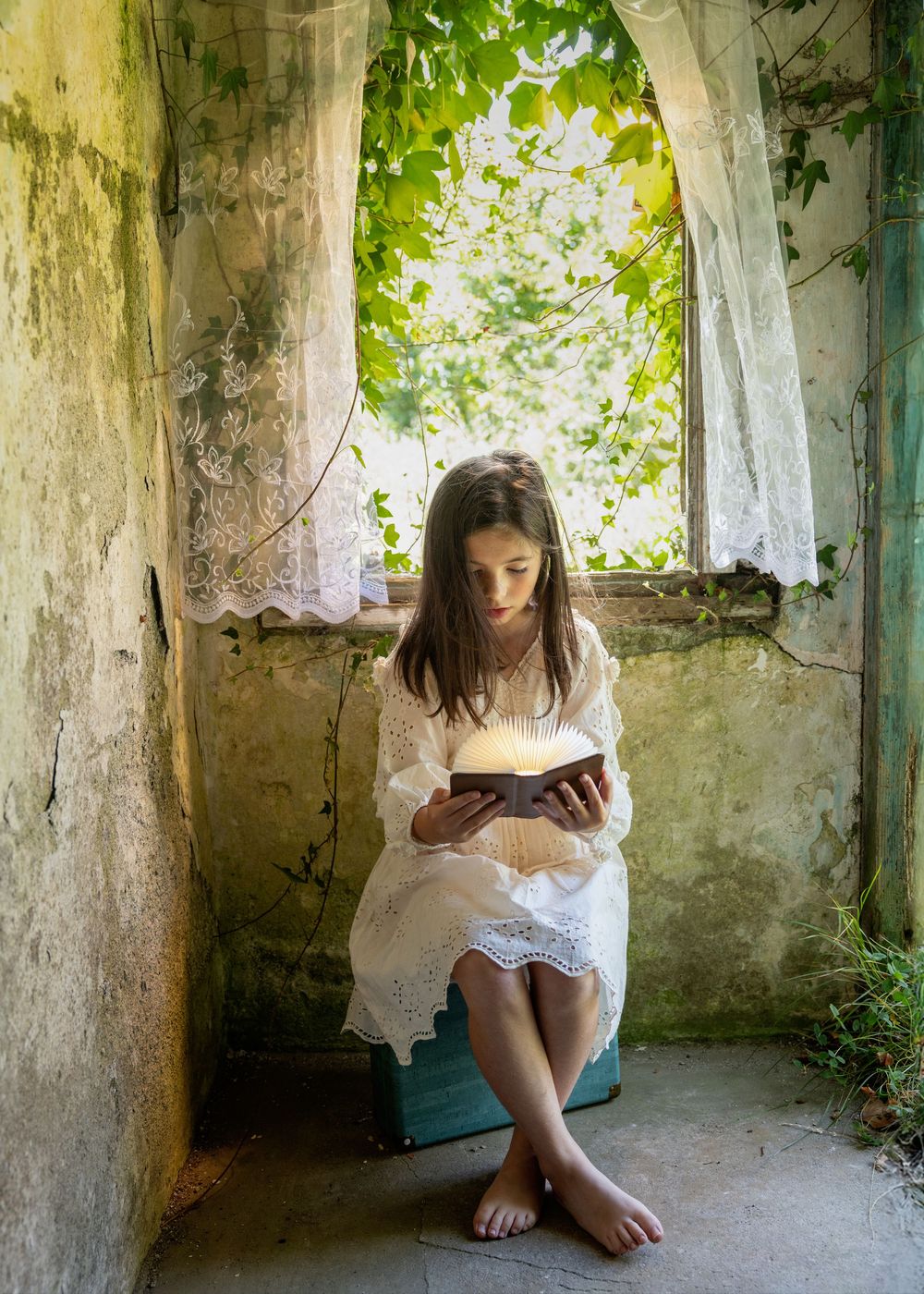 A figure in a white dress reads a book while sitting barefoot in an old stone alcove with vines and natural light.