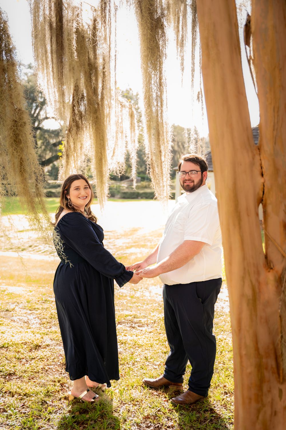 A couple plays peek-a-boo around tree trunks in a sunlit forest during an outdoor photo session.