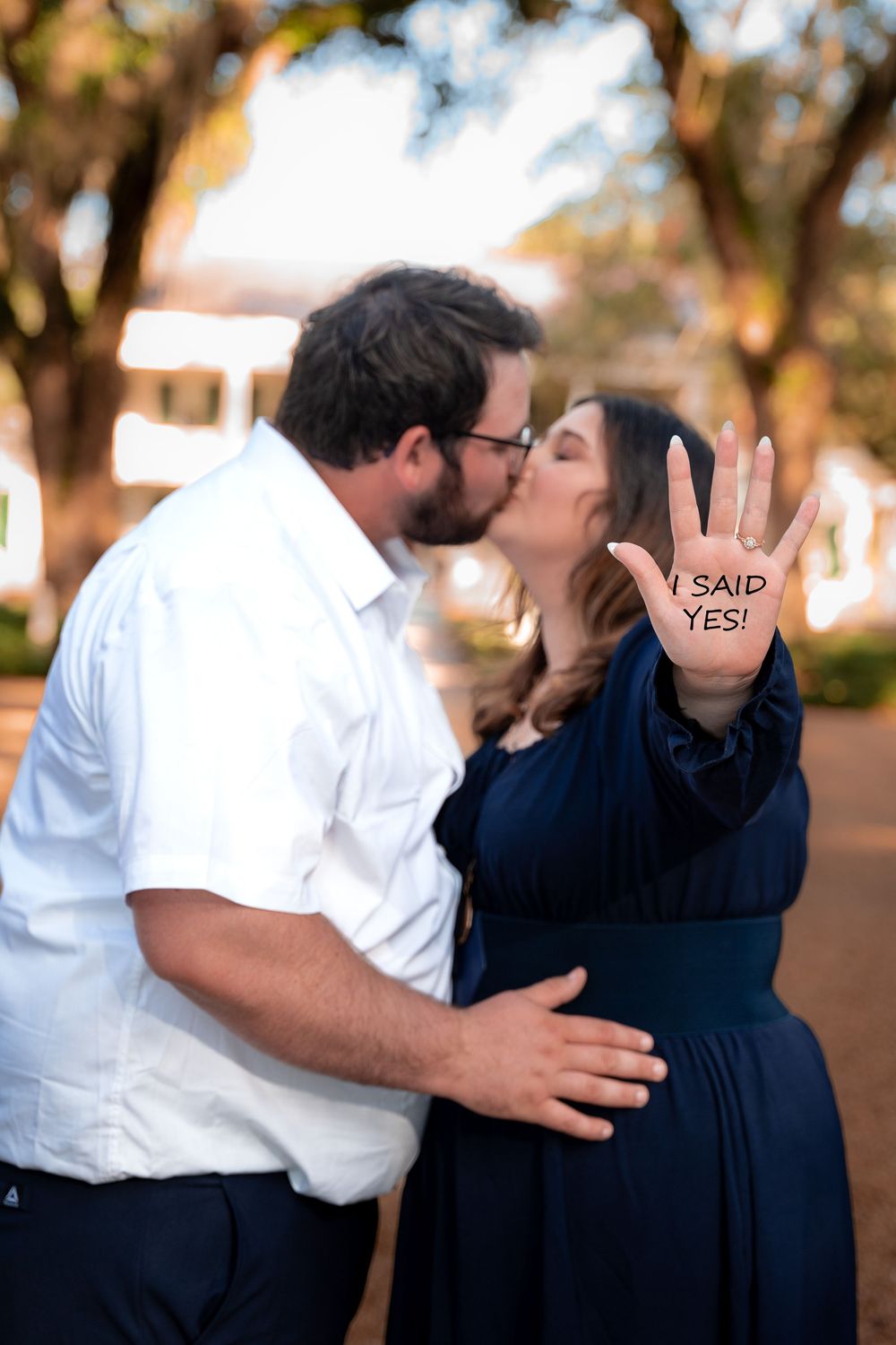 A couple in white and navy blue outfits share romantic kisses in an outdoor setting during a proposal photoshoot.