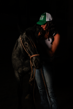 A dark moody photo of a rider in a green trucker hat standing next to a black horse against a black background.