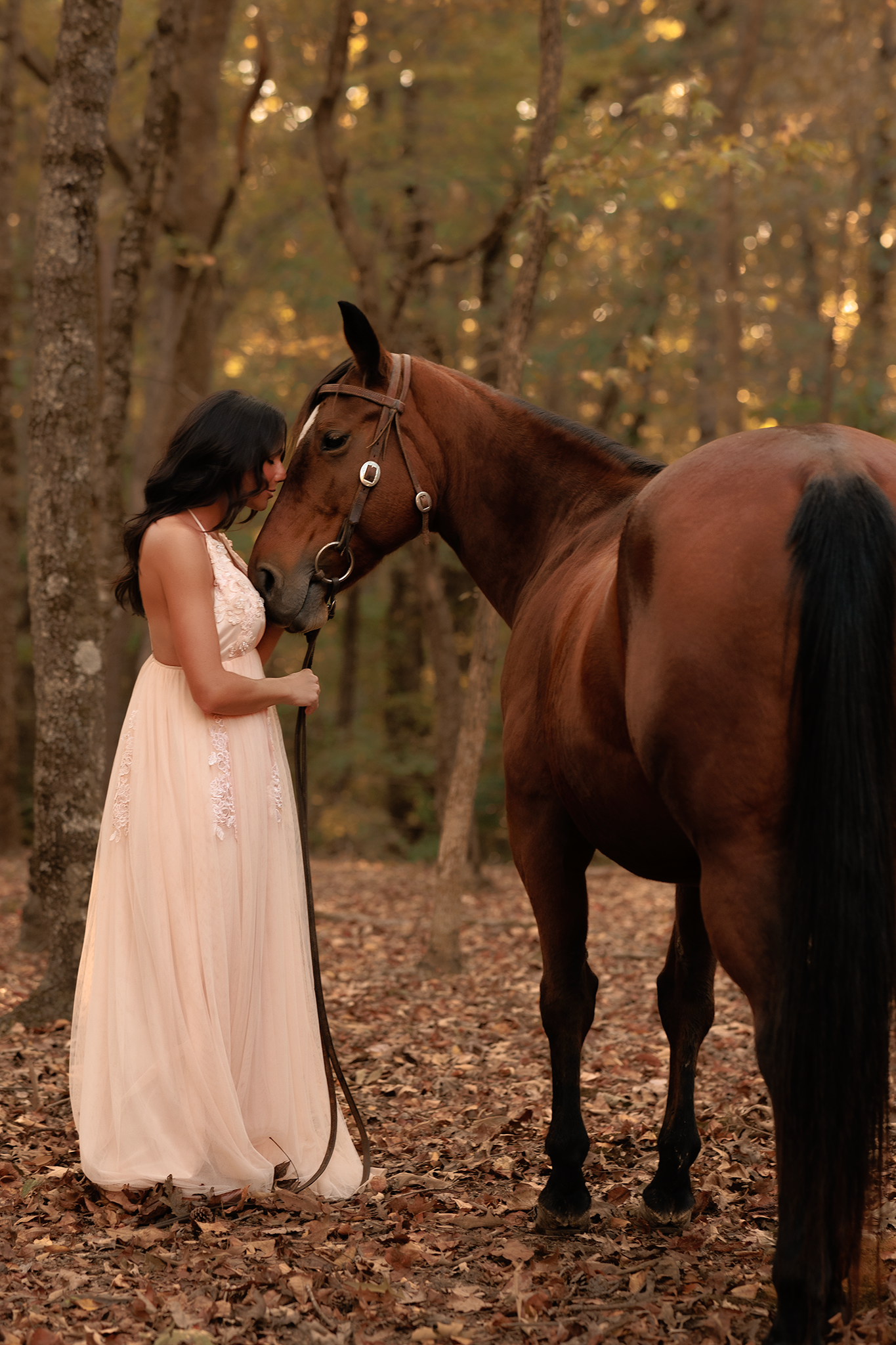 A brown horse and person in a flowing peach gown share a tender moment in an autumn forest setting.